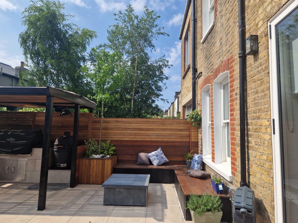 Paved garden with an outdoor kitchen, seating and raised planters. Richmond Garden Design.