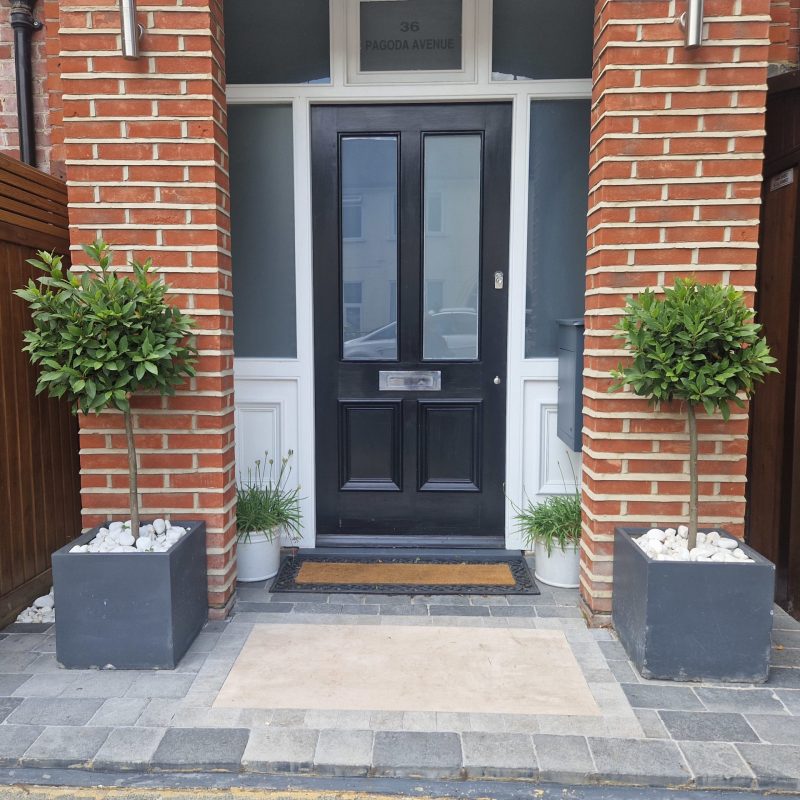 A stone path leading to a wooden front door framed by a beautifully designed patio garden with a medley of green foliage, created by Taylored Gardens.