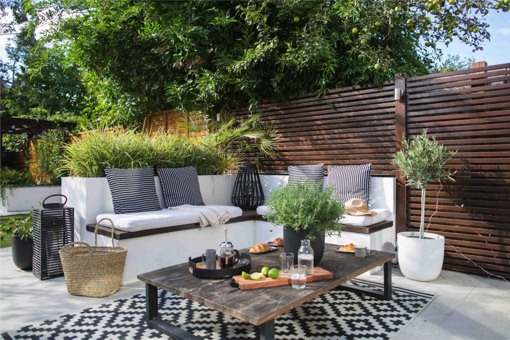 Built-in white corner seating with cushions on a paved patio, with grasses and an olive tree by a slatted fence