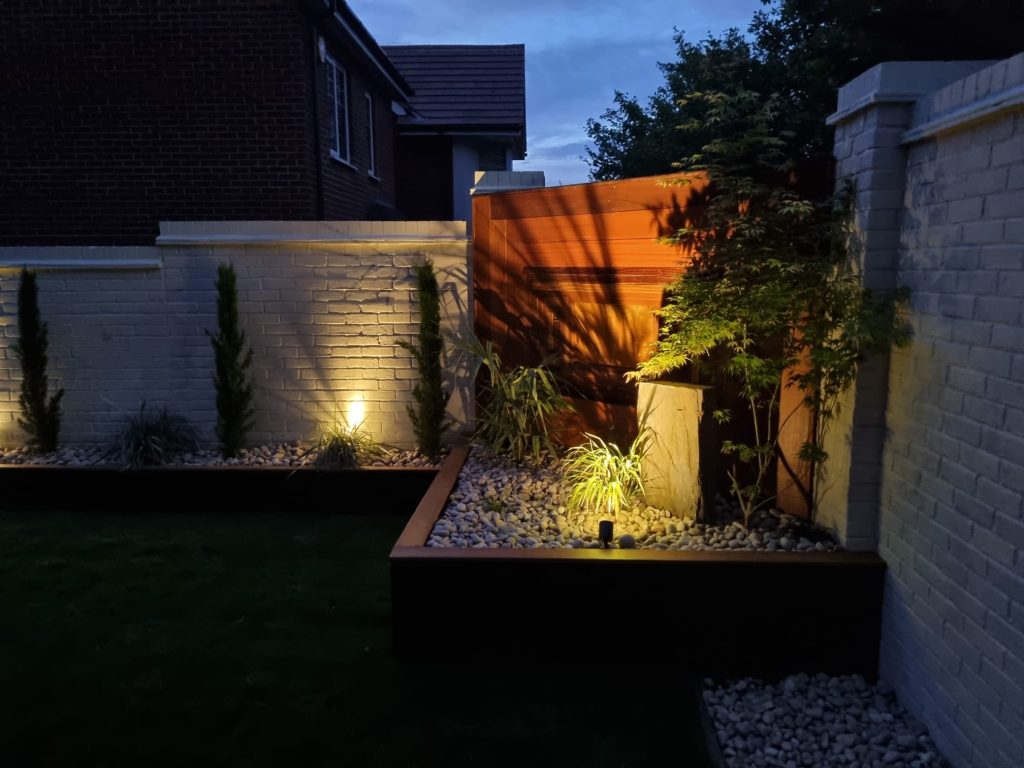 Garden uplighting illuminating raised pebble beds, a Japanese maple and timber screening at dusk