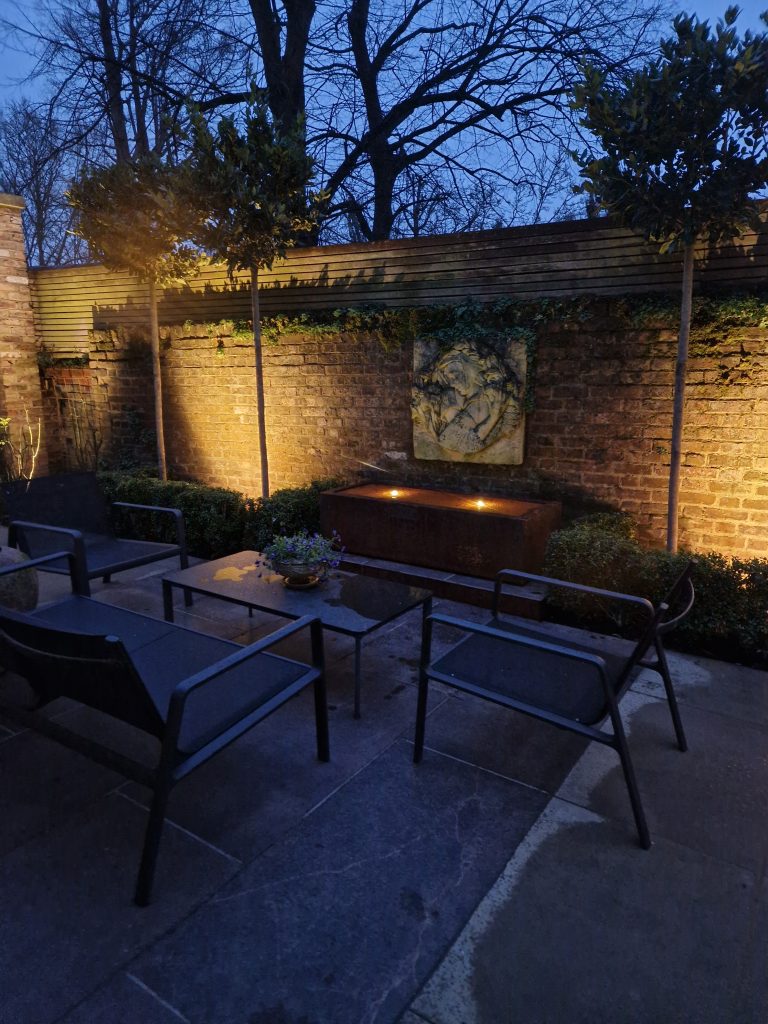 Garden seating area at dusk with warm lighting on a brick wall and timber pergola