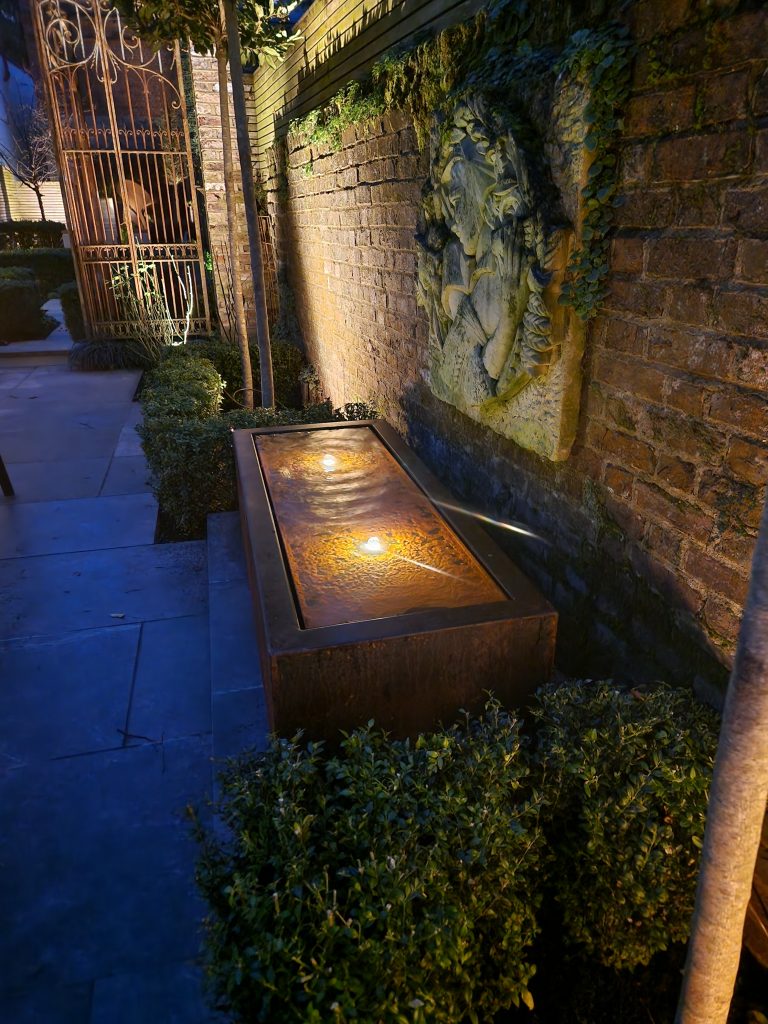 Illuminated corten-steel water feature against a brick wall.