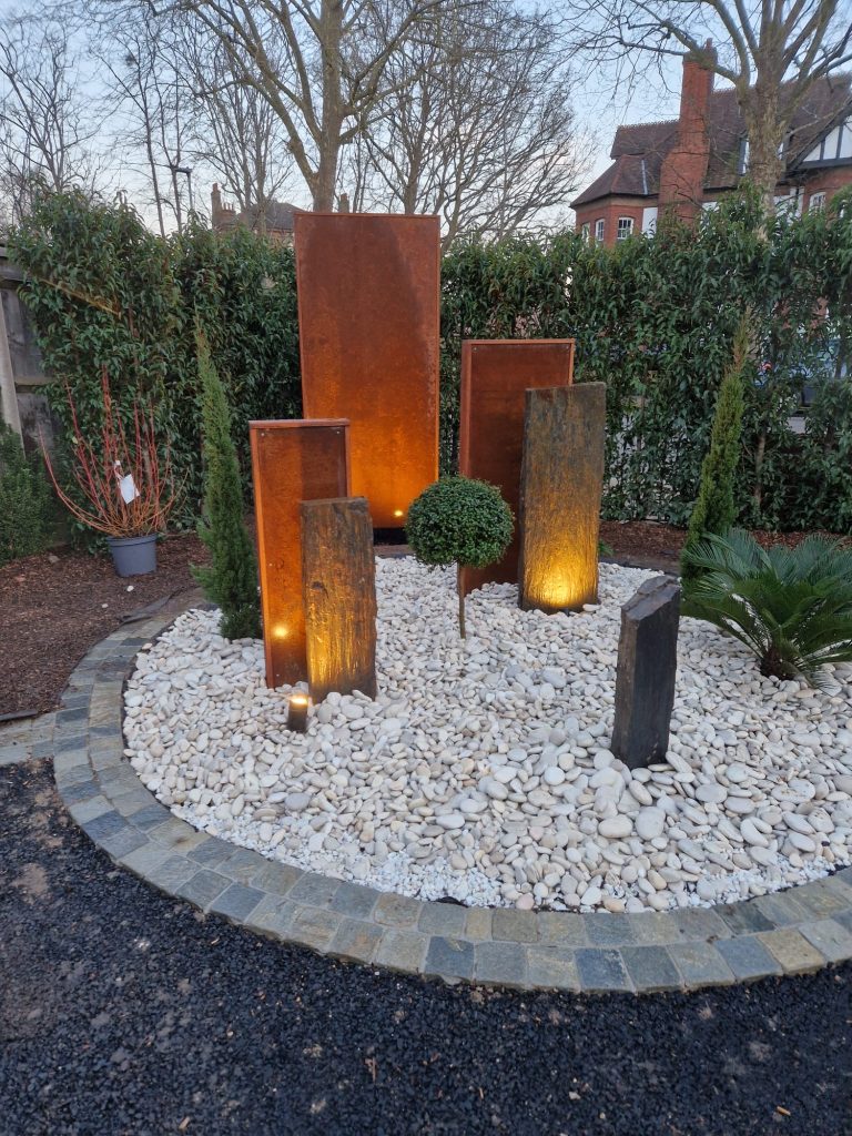 Corten steel panels and slate uprights with uplighting in a white pebble circle with clipped topiary