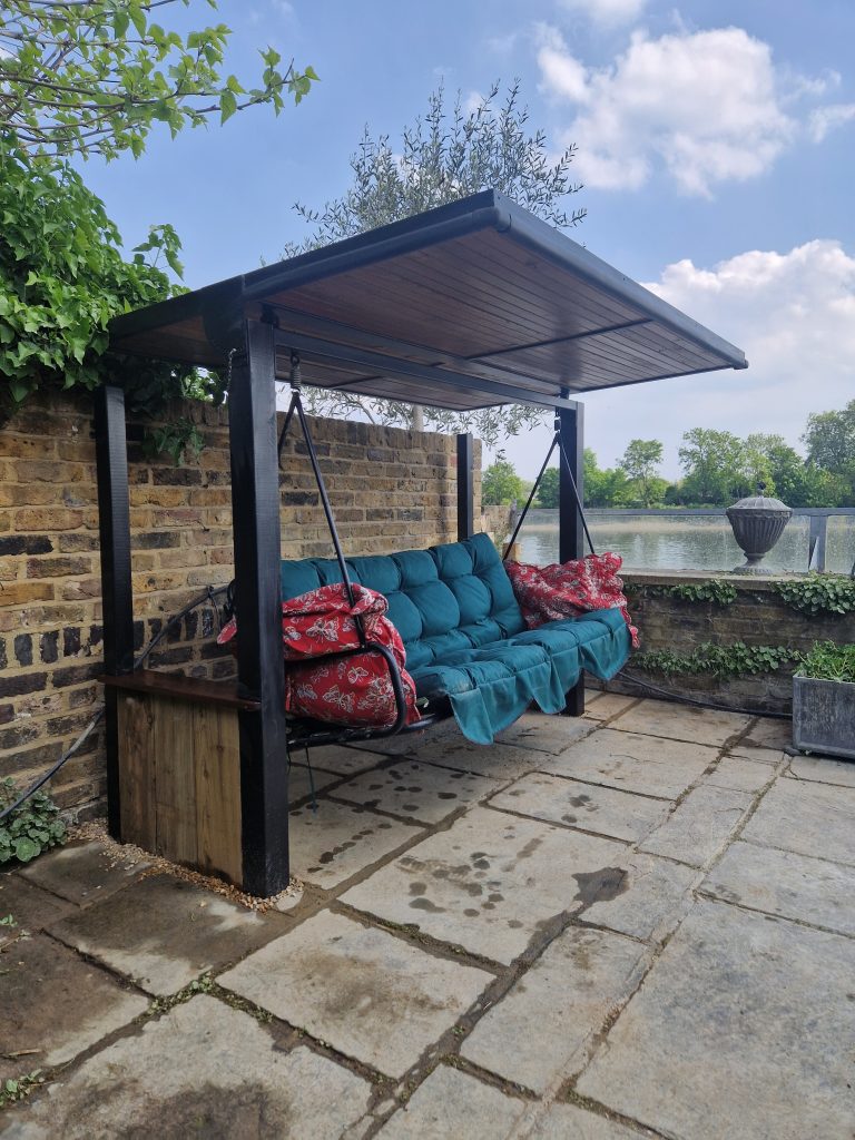 Covered swing seat with teal cushions under a timber canopy on a stone patio by the water