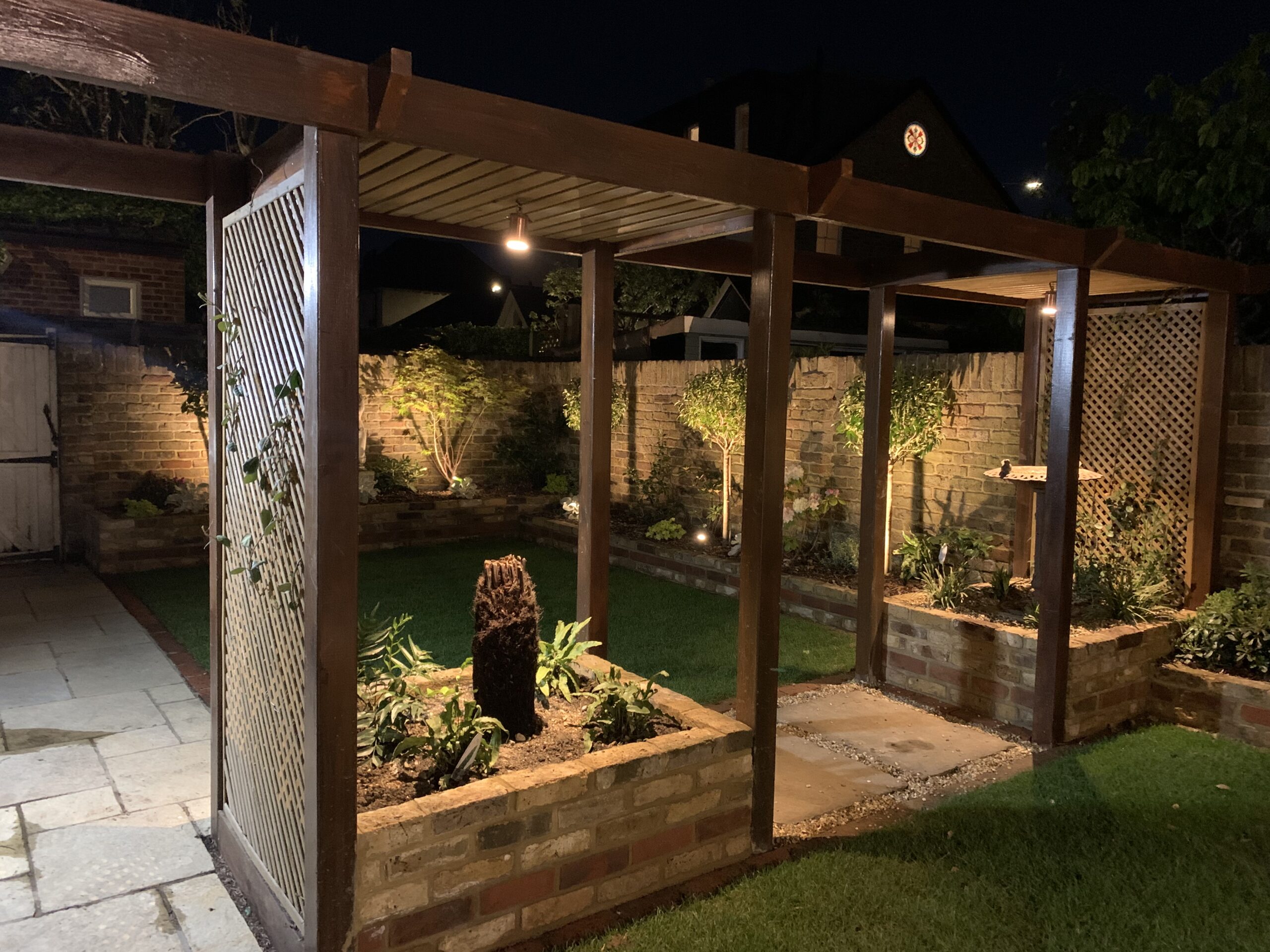Landscaped garden at night with a timber pergola, brick raised beds, uplit trees and lawn